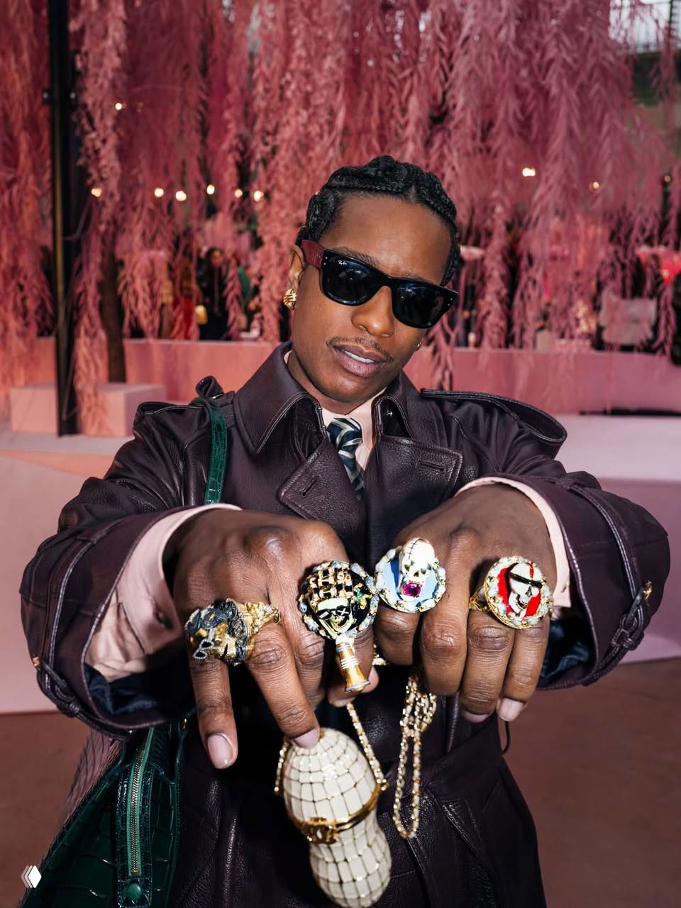 Мужчина в кожаном плаще и солнцезащитных очках демонстрирует крупные кольца и аксессуары на фоне розового декора кутюрного показа.