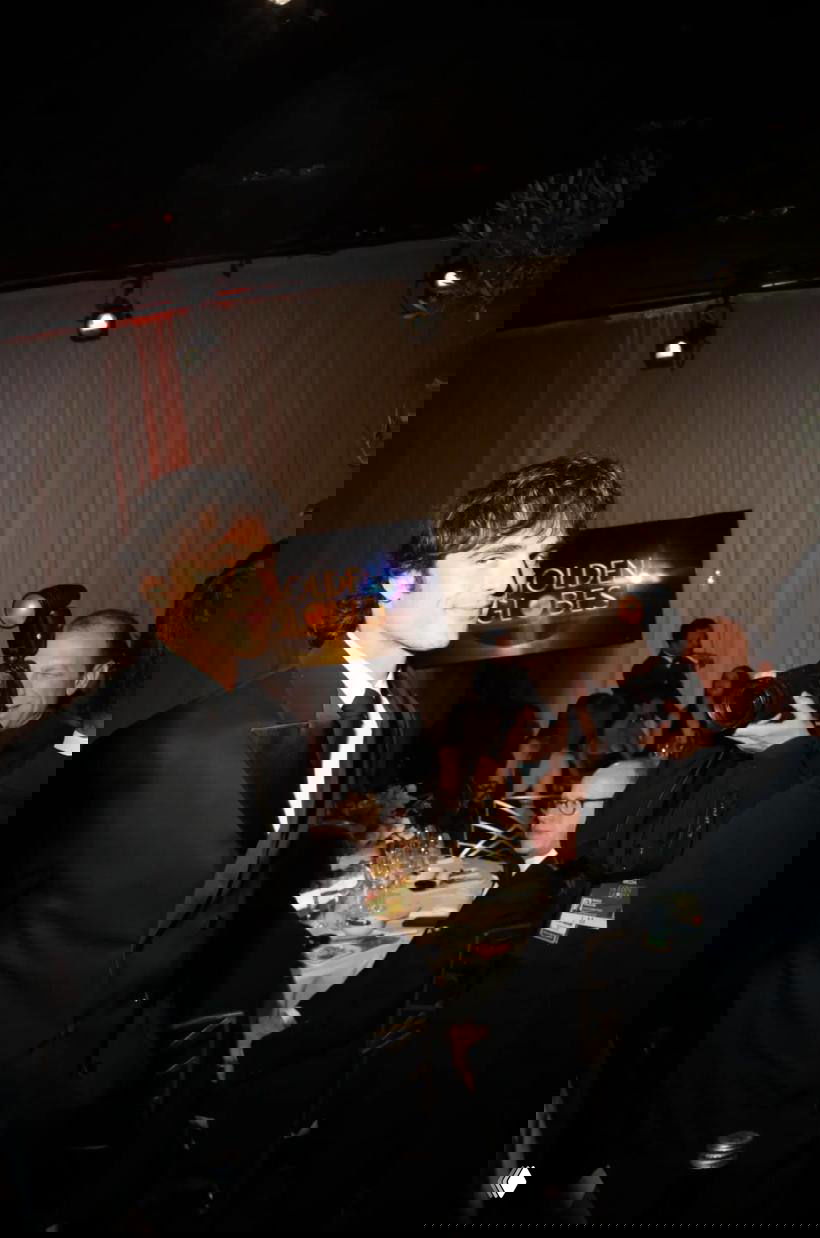 Мужчина в классическом чёрном костюме идёт по залу на приёме Golden Globes, фотографы делают снимки, видно столы и приглашённых гостей.