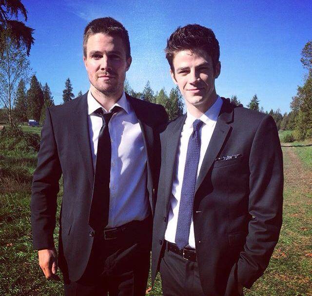 Два мужчины в классических костюмах стоят на зелёной поляне и улыбаются — неформальный кадр за кадром со съёмок или мероприятия.