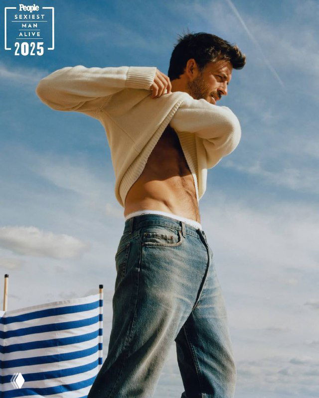 Джонатан Бейли снимает свитер на открытом воздухе в кадре фотосессии People 2025: видно плечо, торс, джинсы и небесный фон.