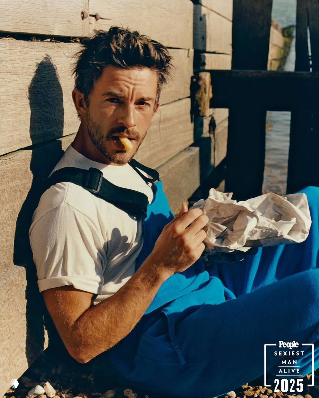 Портрет Джонатана Бейли у деревянного пирса: актёр в рабочем комбинезоне сидит на берегу с закуской, лёгкая небритость и тёплые тона снимка.