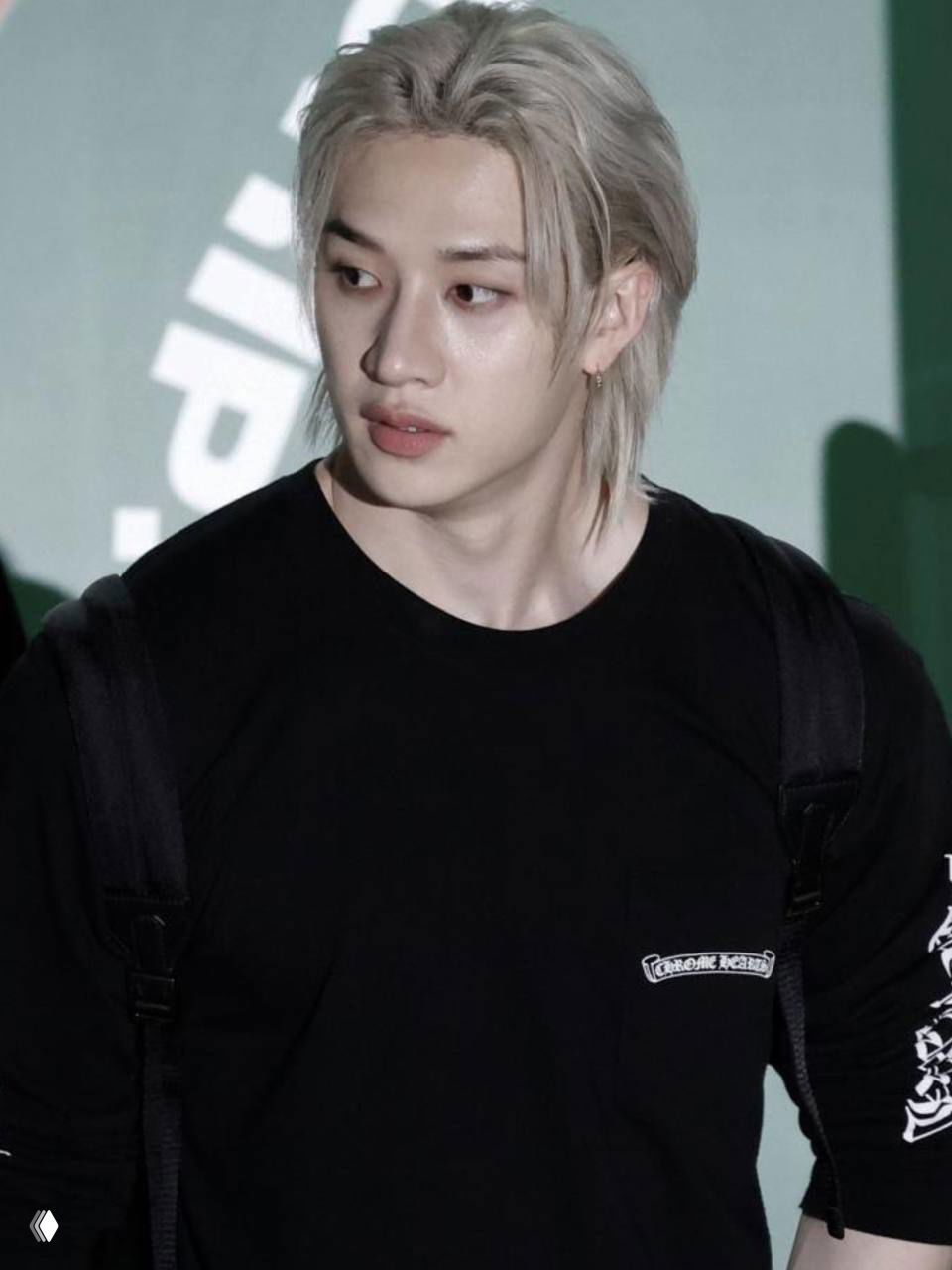 Крупный план молодого человека в чёрной футболке и рюкзаке, светлая причёска и серьга, фотография в уличном/фан-стиле; личность не указывается.