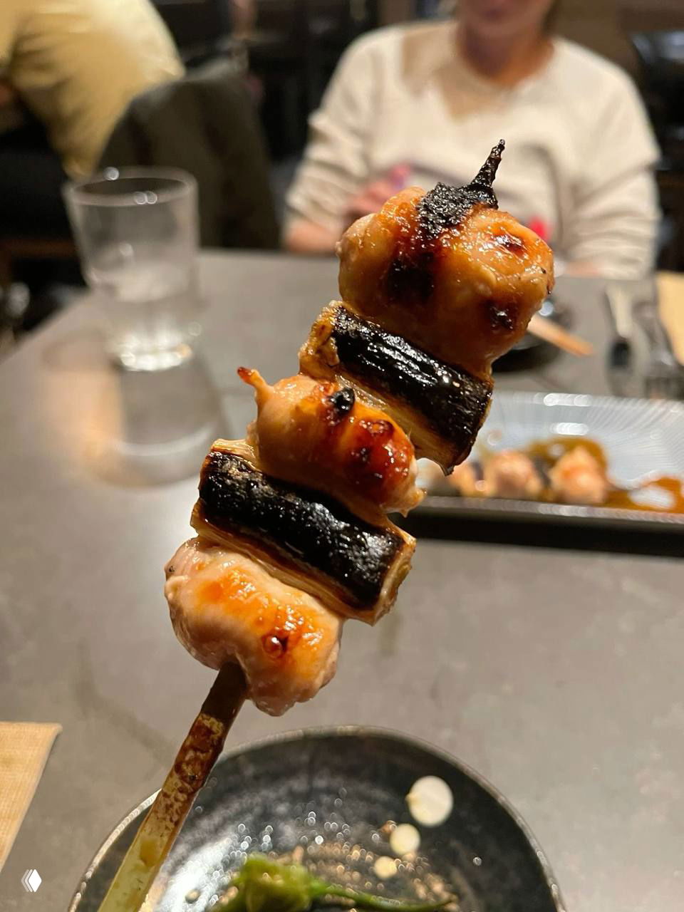 Крупный план якитори негима на деревянной шпажке: поджаренные кусочки курицы и лук-порей на фоне столика и размытых посетителей ресторана.