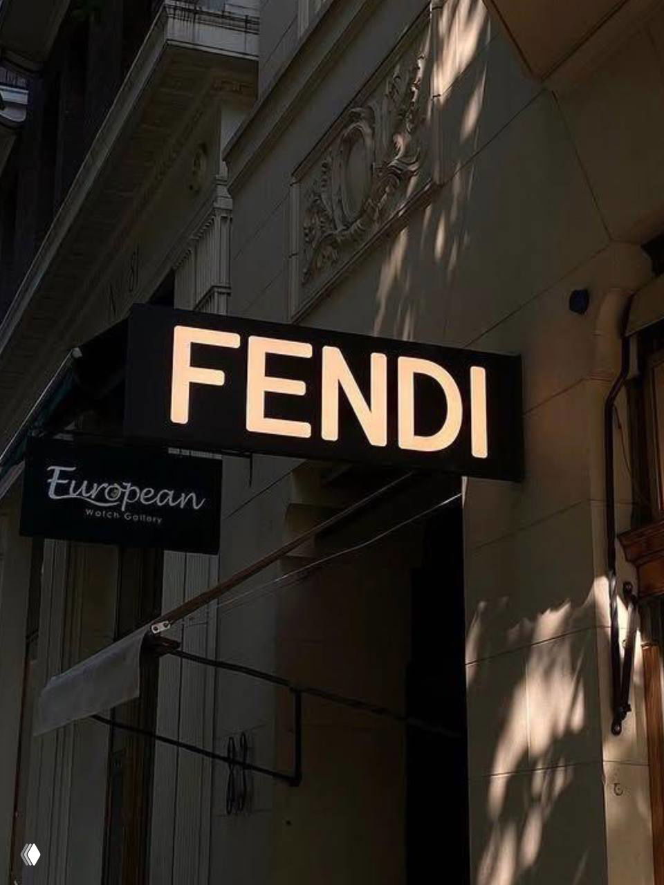 Вывеска FENDI на фасаде здания с тёплым боковым освещением и архитектурными тенями, городской контекст вокруг витрины.