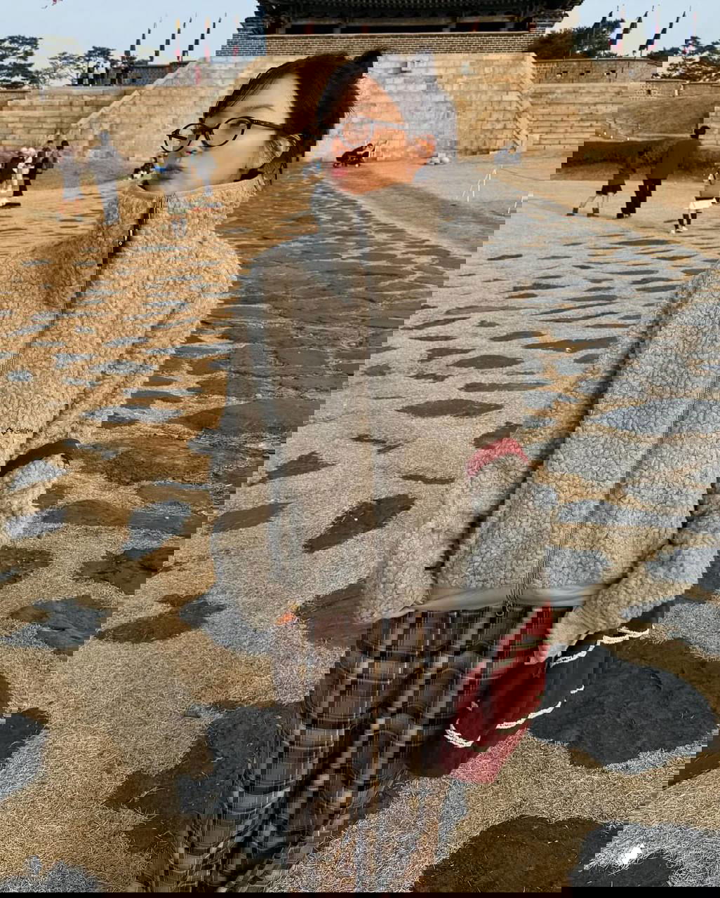 Молодая женщина на открытом воздухе в серой тёплой пушистой куртке на замке и в очках, образ в стиле уличной корейской моды на фоне каменной площади.