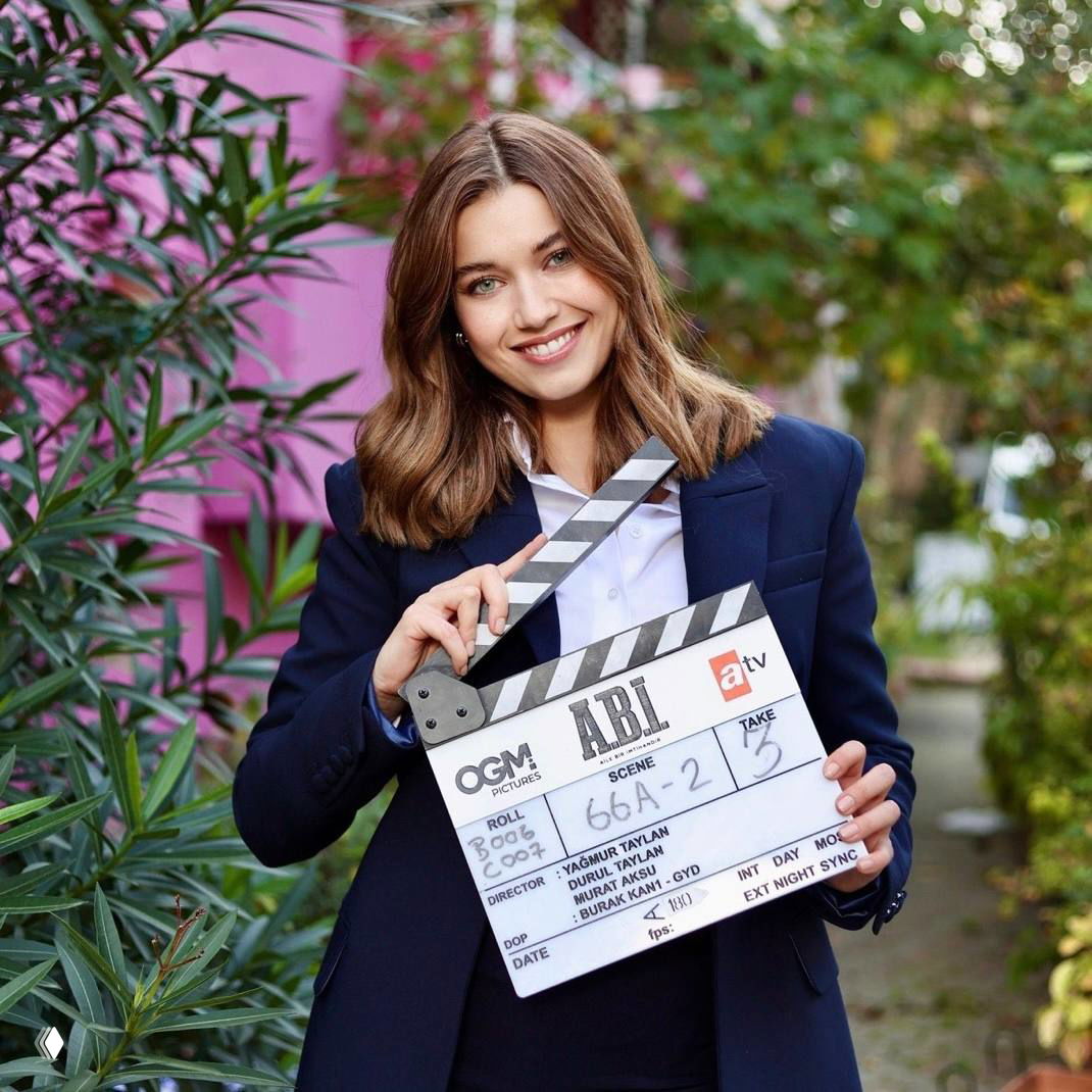 Улыбающаяся молодая женщина в жакете держит хлопушку для киносъёмки на фоне зелёных растений и яркой стены, кадр со съёмочной площадки.