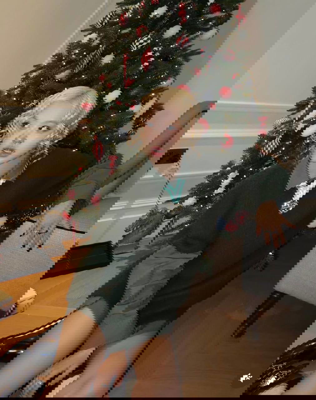 Айдол в зелёном свитере и светлой причёске позирует на фоне украшенной ёлки; расслабленный кэжуал-образ с декоративной брошью и чулками.