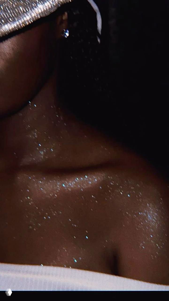 Ночной кадр декольте и плеч с рассыпанным блеском: мелкие светящиеся частицы на тёмной коже создают эффект звёздной пыли.