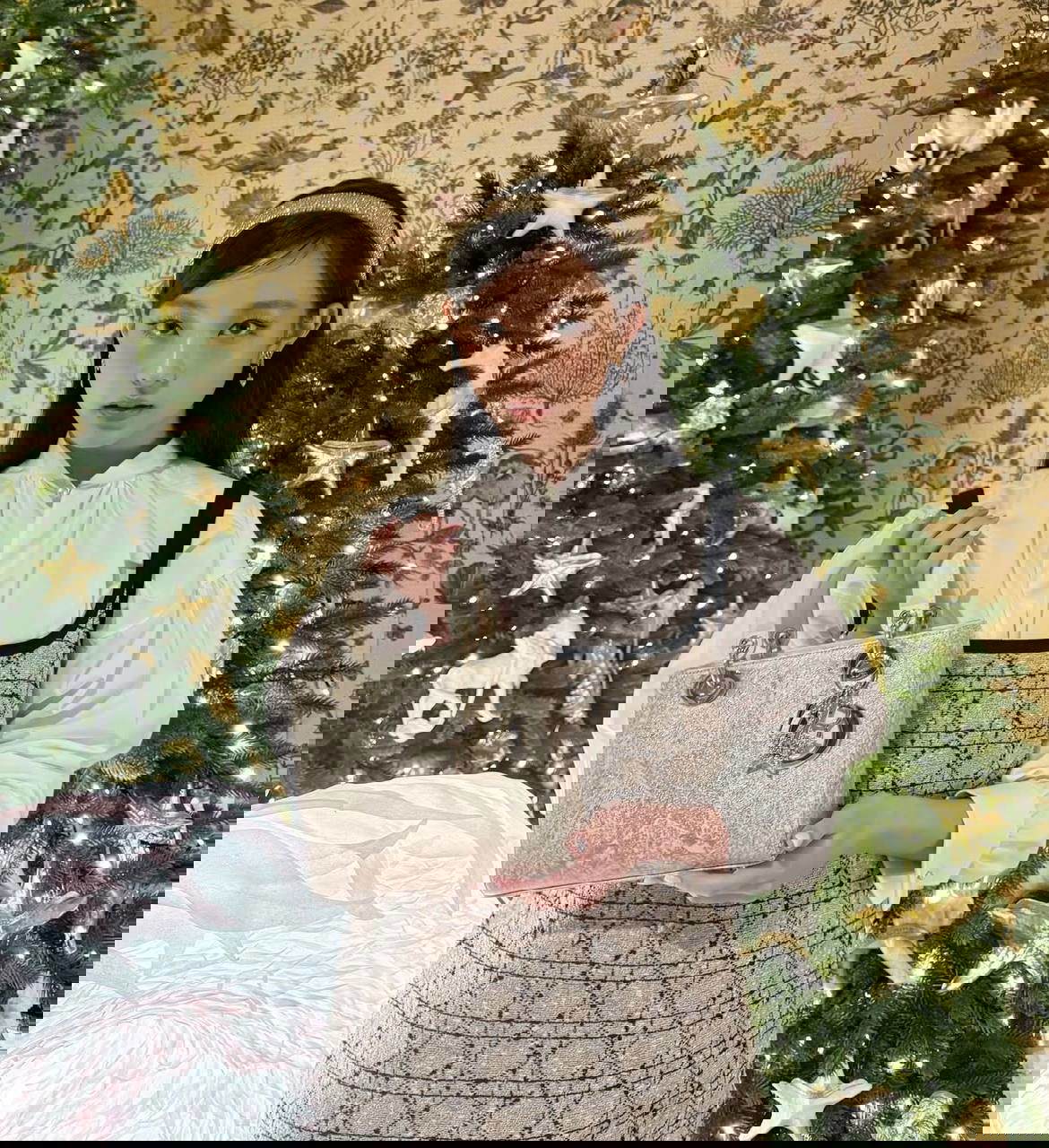 Айдол в светлом платье и повязке держит блестящую сумку на фоне двух рождественских ёлок; образ в романтической праздничной эстетике.