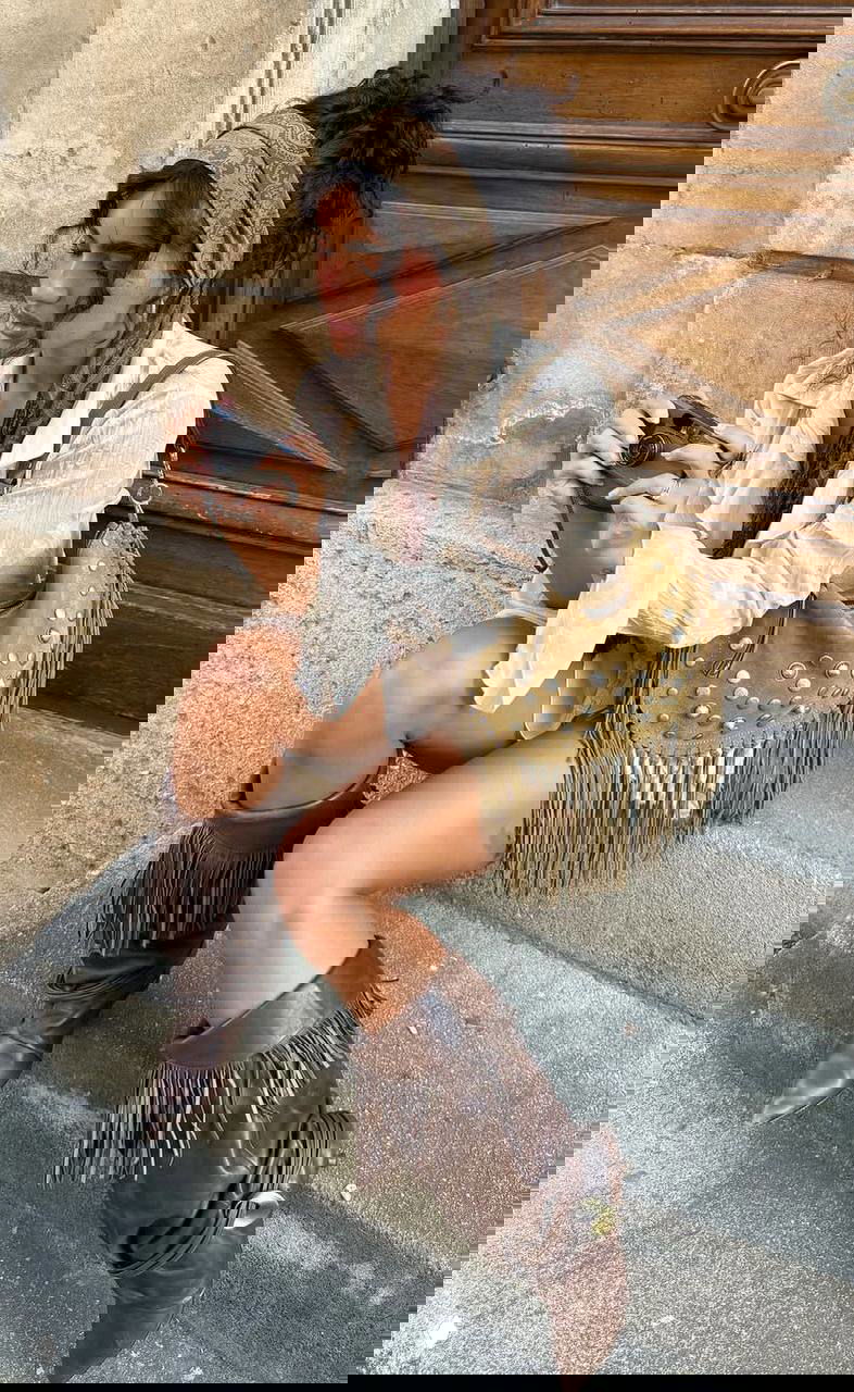 Уличный снимок девушки, сидящей на бордюре в коричневых высоких сапогах с бахромой и сумкой в стиле бохо — комплект в теплых тонах и фактуре.