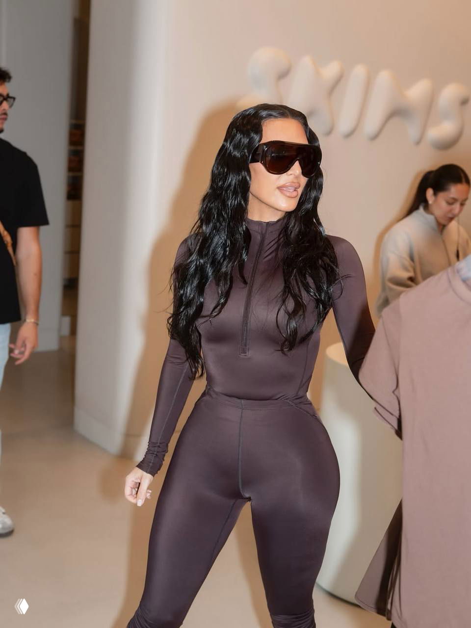 Женщина в облегающем тёмном костюме и больших солнцезащитных очках идёт по интерьеру бутика SKIMS; на заднем плане посетители и экспозиция.