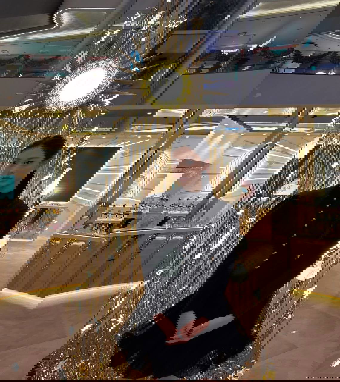 Айдол в чёрном платье позирует у золотой праздничной инсталляции в магазине или бутике; вечерний образ с минималистичным макияжем и аксессуарами.