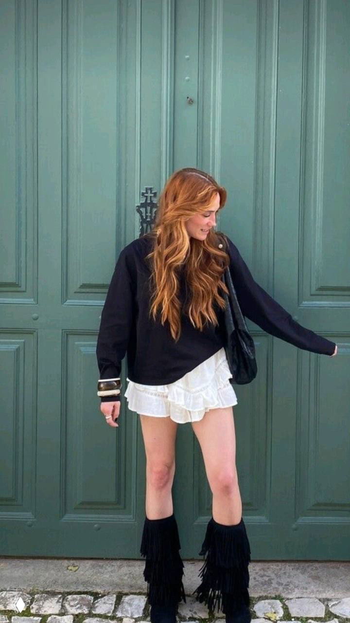 Полнотелая фотография девушки у двери в высоких черных сапогах с несколькими ярусами бахромы — легкий контраст с мини‑юбкой и свитером.