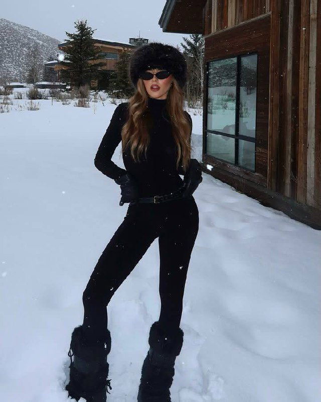 Женщина в черном облегающем кэтсьюте, меховой шапке и солнцезащитных очках позирует на фоне деревянного шале и снега, элегантный спортивный образ.