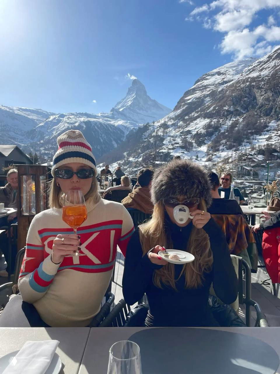 Две девушки за столиком на террасе у гор, одна держит аперитив, другая — чашку кофе; на заднем плане заснеженная вершина Маттерхорна, атмосферный горный отдых.