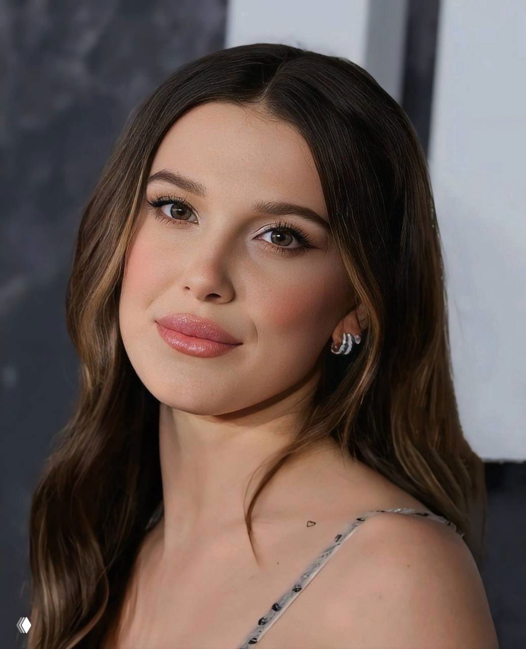 Крупный план лица Милли Бобби Браун на мероприятии: аккуратный макияж, распущенные волосы и мягкий нейтральный фон, портретный кадр.