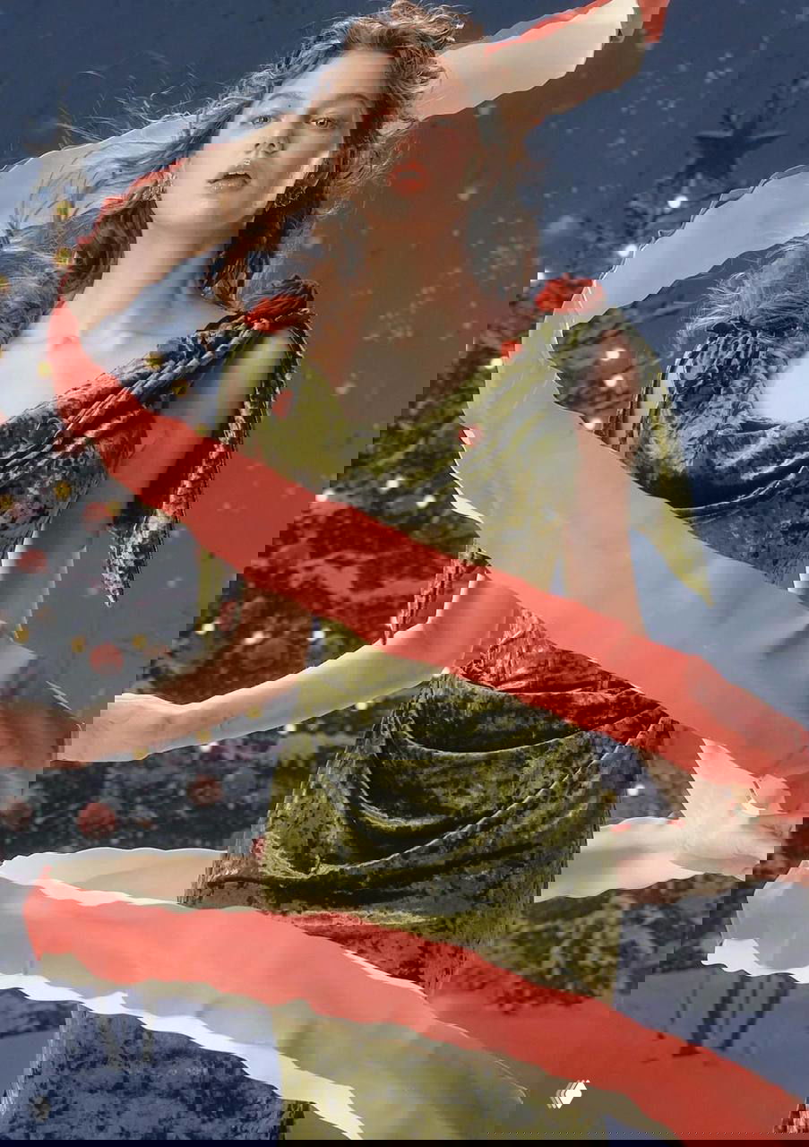 Модель в зелёном бархатном платье в сказочном стиле, украшенном цветами и лентами, на фоне заснеженной рождественской ёлки и огней.