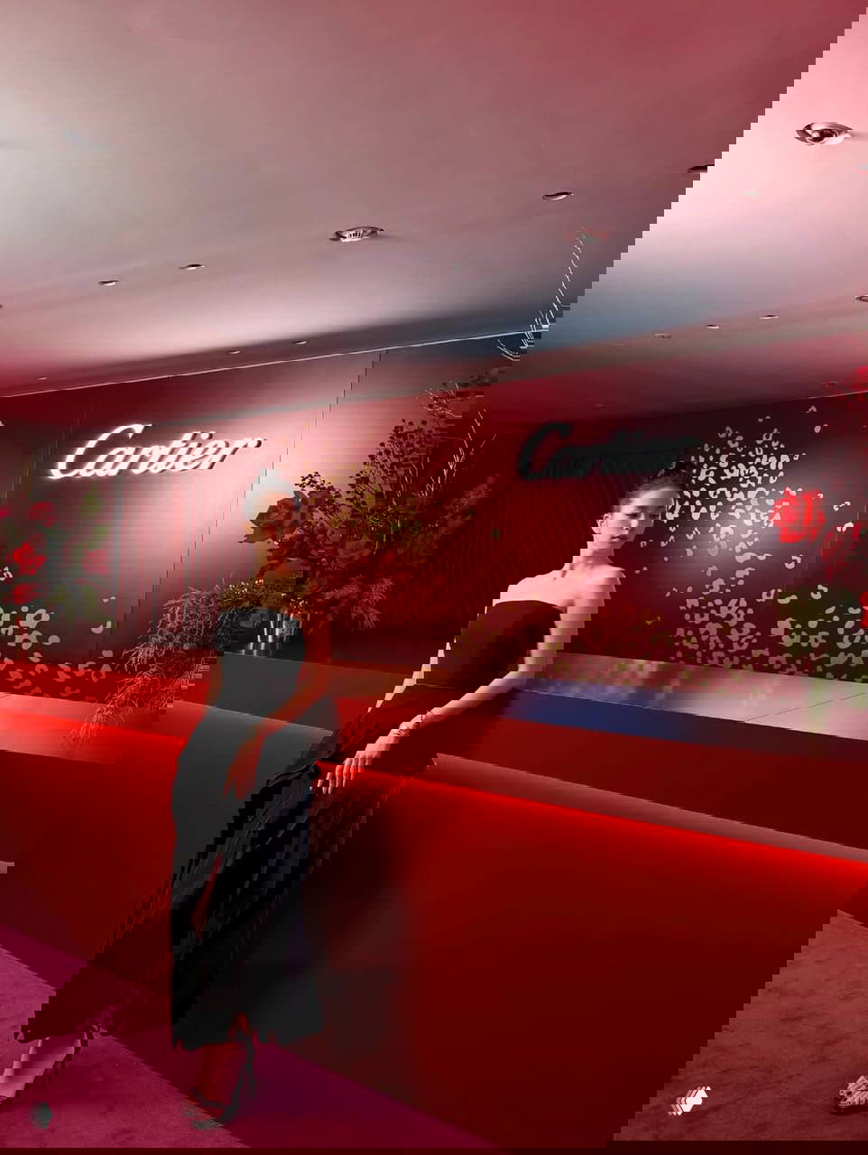 Джису стоит у длинной красной сцены с надписью Cartier, в длинном чёрном платье и туфлях на каблуке, минималистичный праздничный интерьер.