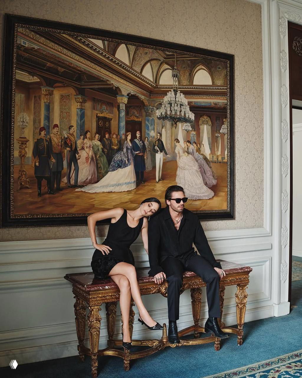 Зейнеп Оймак и Таро Эмир Текин в элегантных образах сидят на резном столе на фоне большой исторической картины; интерьер передаёт эстетику old money.