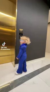 Двухлетний ребёнок с длинными светлыми волосами и в синем костюме идёт по холлу у лифта, вертикальный кадр, видно пол и стену