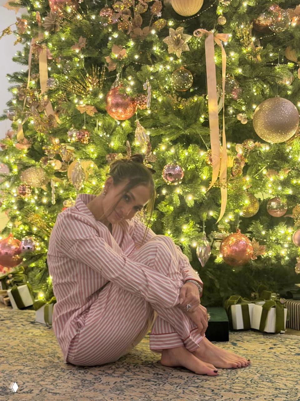 Дженнифер Лопес в полосатой пижаме сидит у большой наряженной ёлки, согнув ноги, вокруг мерцают огни и рождественские украшения, мягкий интерьер.