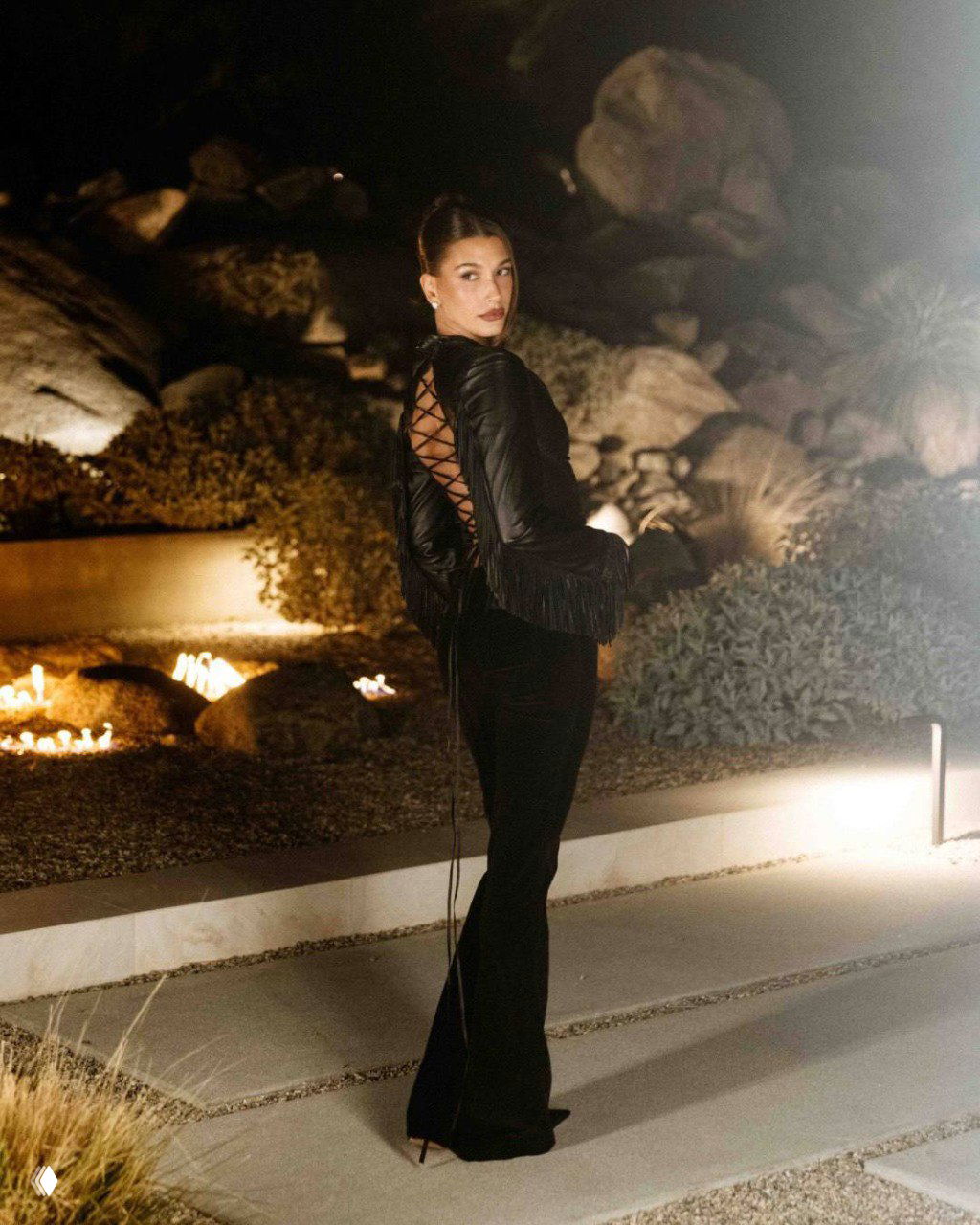 Хейли Бибер на открытой терассе вечером в полный рост: черная кожаная комбинация с декоративной шнуровкой и бахромой, теплые огни на заднем плане.