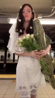 Девушка в белом свадебном платье и фате держит букет и большого аллигатора на фоне станции — кадр из вирусного ролика к Хэллоуину.