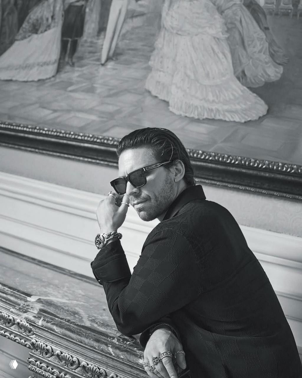 Портрет Таро Эмир Текина в чёрно‑белой гамме: актёр в очках и украшениях опирается на антикварную мебель, подчёркнуты детали образа и аксессуары.