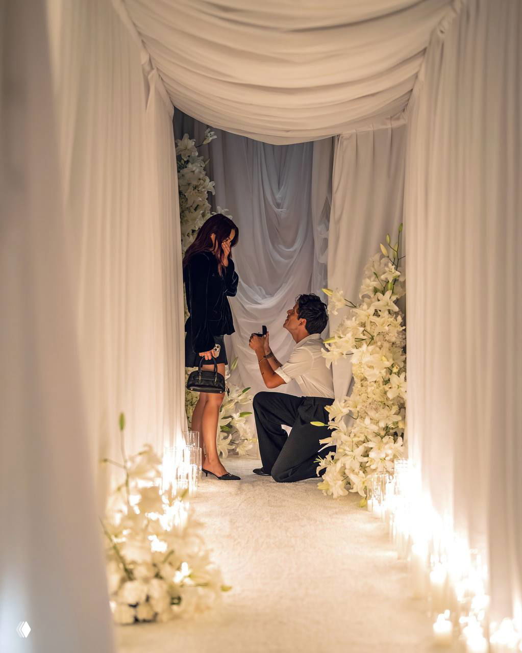 Камила Мендes в чёрном платье, Руди Манкузо опустился на колено с коробочкой-кольцом в белом коридоре, украшенном цветами