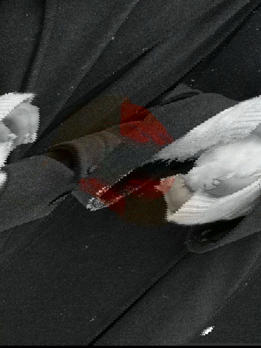 Руки в белых вязаных митенках держат снежок на фоне тёмного пальто — акцент на зимних аксессуарах, текстурах и уютной стилистике.