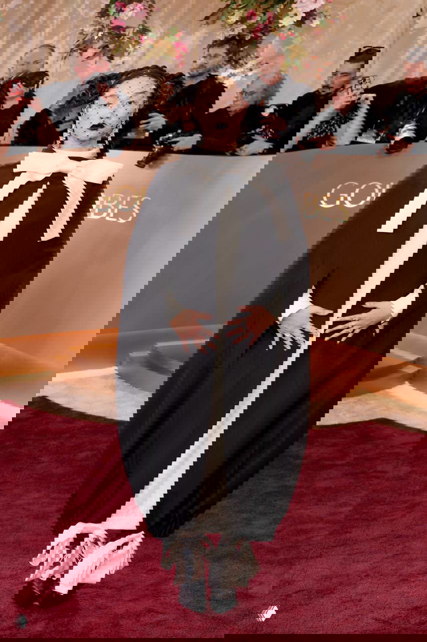 Фигура в чёрной накидке с крупным белым бантом и вуалью на лице позирует на красной дорожке на фоне логотипа мероприятия и камер.