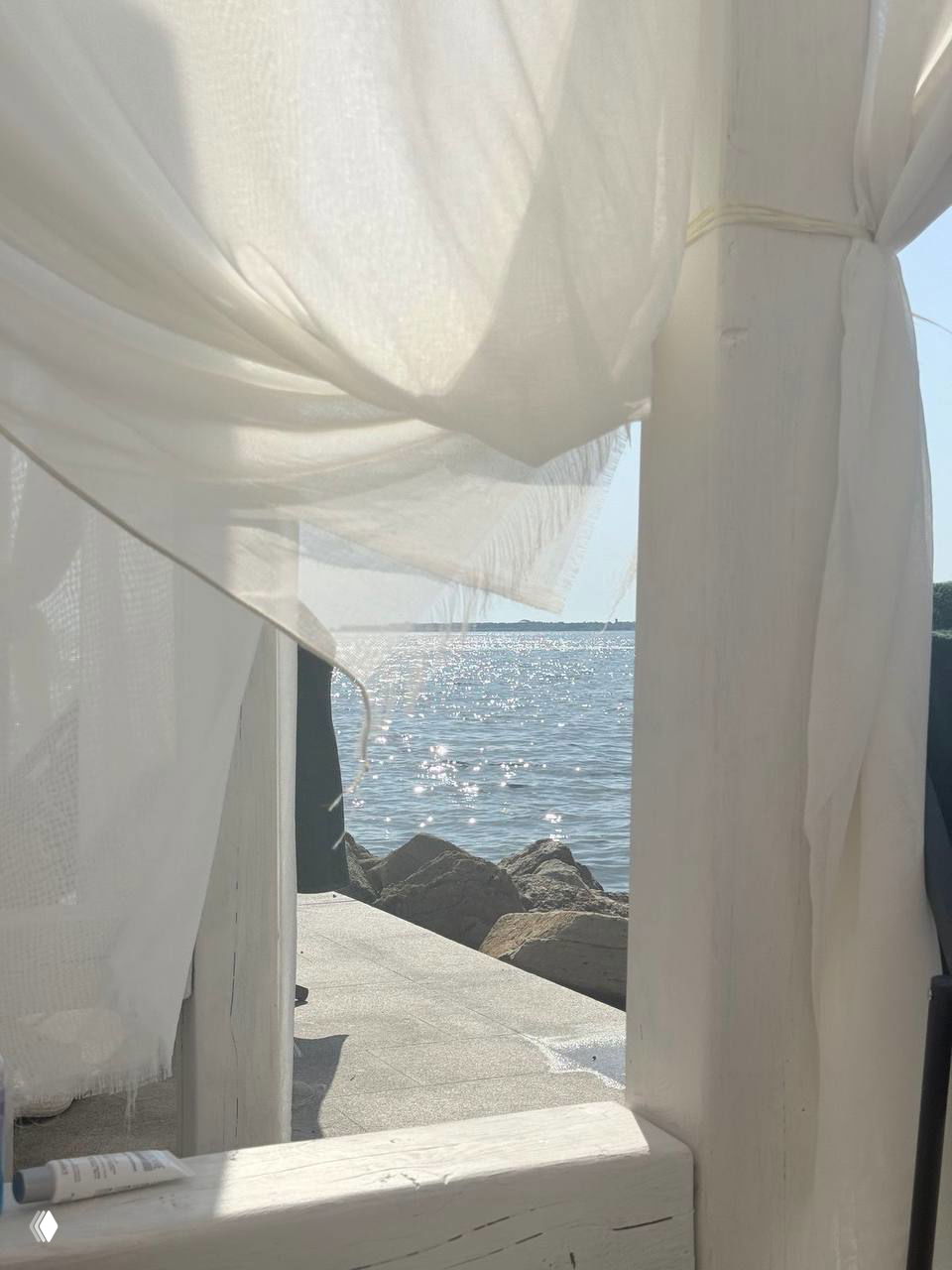 Пляжный кадр: белая прозрачная занавеска развевается у воды, виднеются камни и солнечные блики на море — светлая атмосфера образа.