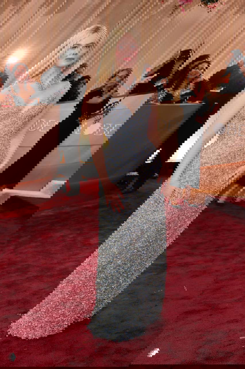 Женщина в блестящем светло-голубом платье с аккуратным декольте на красной дорожке, вокруг — фотографы и зрители в холле мероприятия.