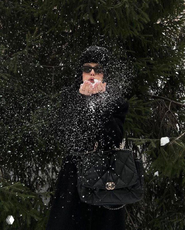 Девушка в тёмном пальто и очках разбрасывает снег руками перед хвойным фоном — динамичный зимний кадр с чёрной сумкой-«квинтэссенцией» образа.