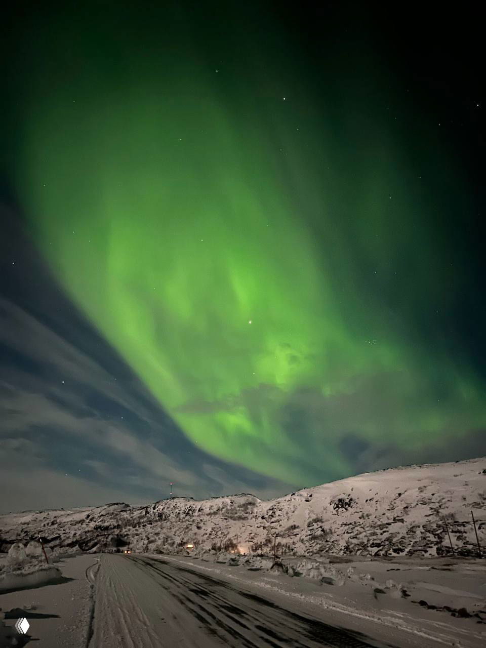 Широкое зелёное северное сияние, растянувшееся по ночному небу над заснеженной дорогой и холмами; виден зимний пейзаж и звёзды.