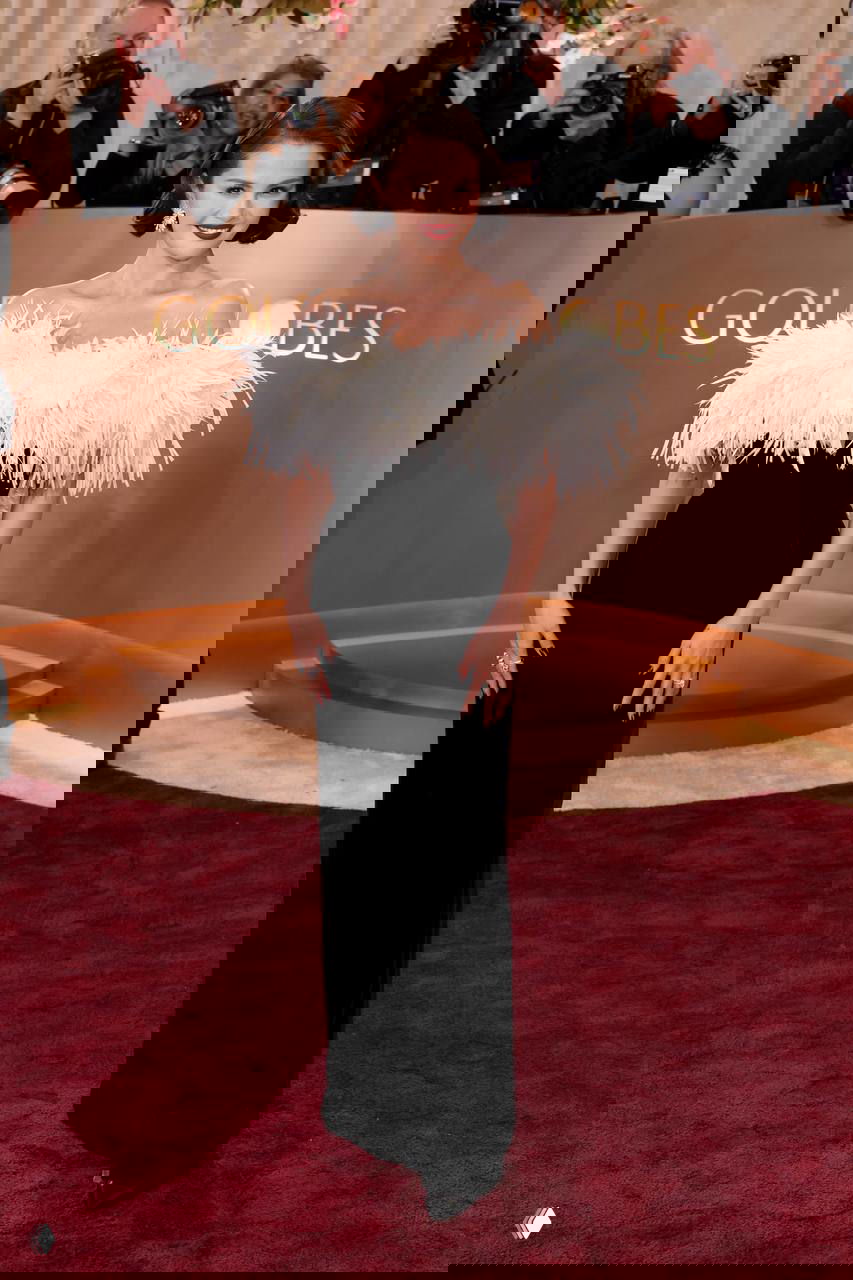 Женщина в чёрном платье с белой перьевой отделкой по линии плеч, улыбается на красной дорожке перед линией фотографов и логотипом мероприятия.