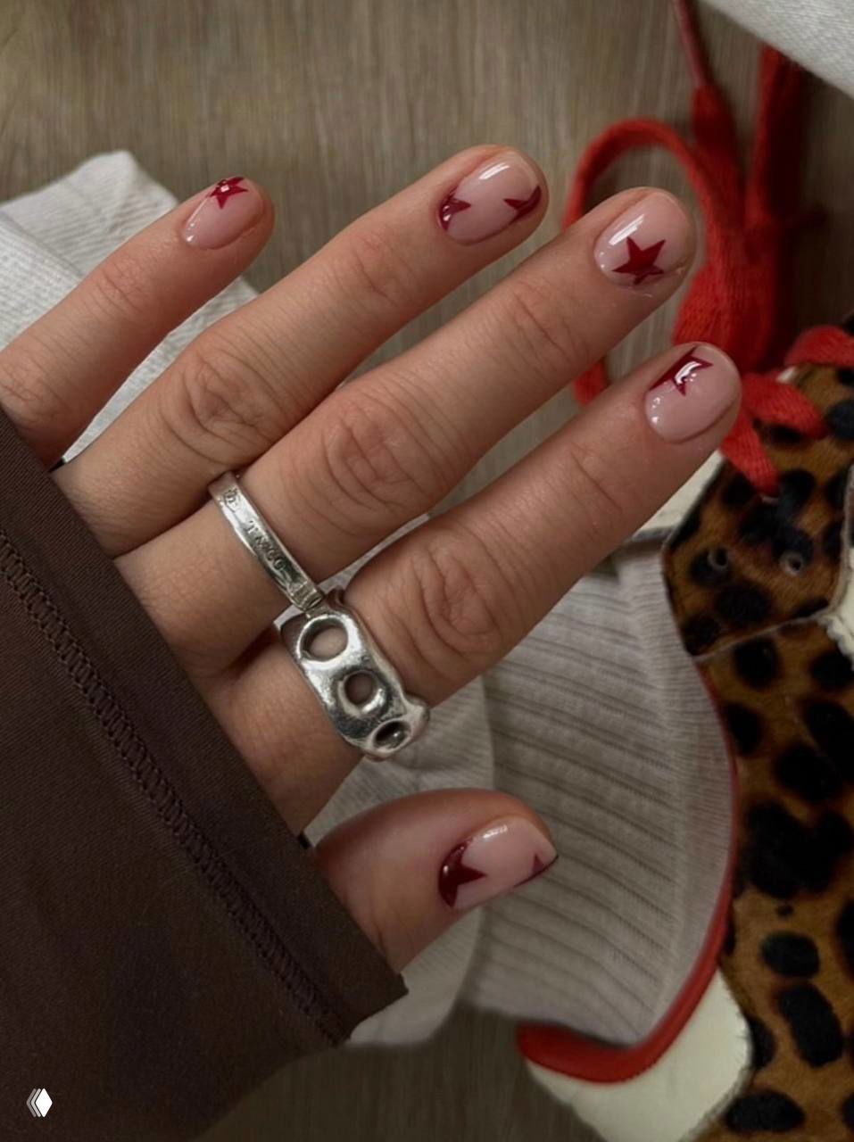 Крупный кадр руки с короткими нюдовыми ногтями и маленькими бордовыми звёздами у края пластины, видны серебряные кольца и текстиль.