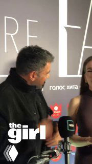 Крупный план Эмре Алтуга в профиль во время интервью на мероприятии; виден микрофон, пресс‑стена и водяной знак «the girl».