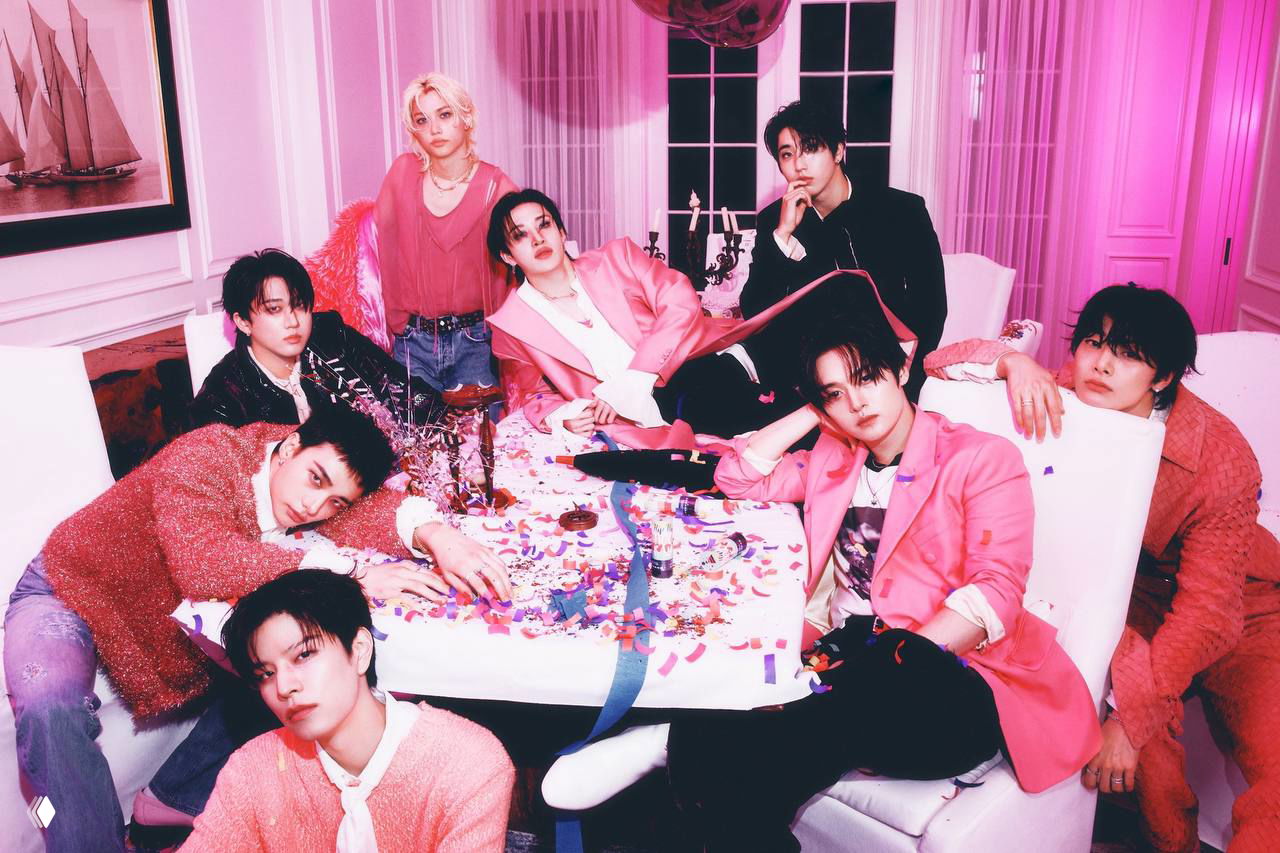 Группа Stray Kids в розовой палитре на промо‑фото: участники вокруг стола с конфетти, праздничная стилизация и яркое освещение в комнате.