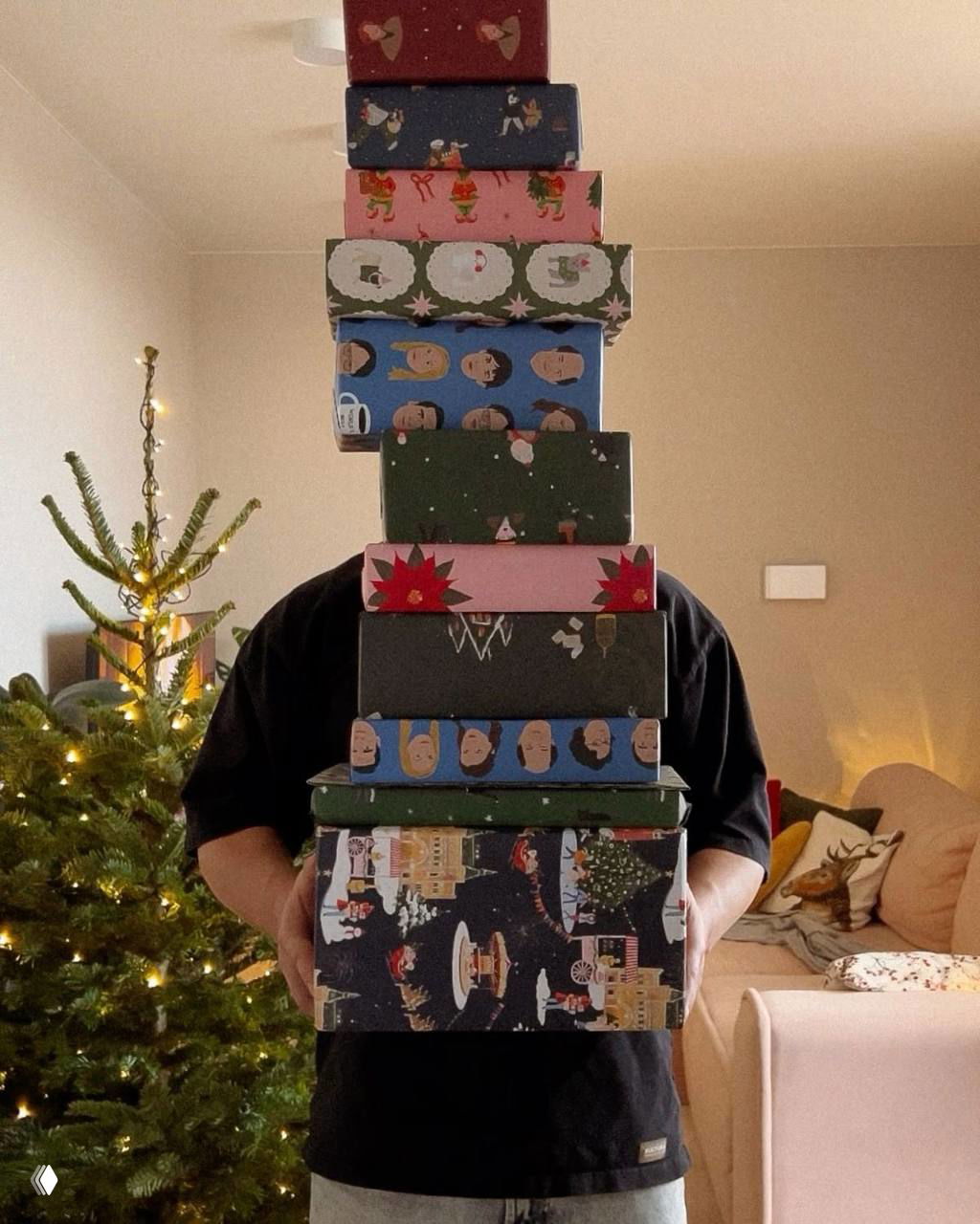 Человек держит высокую стопку красиво упакованных подарочных коробок на фоне украшенной ёлки и гостиной — идея для упаковки множества подарков.