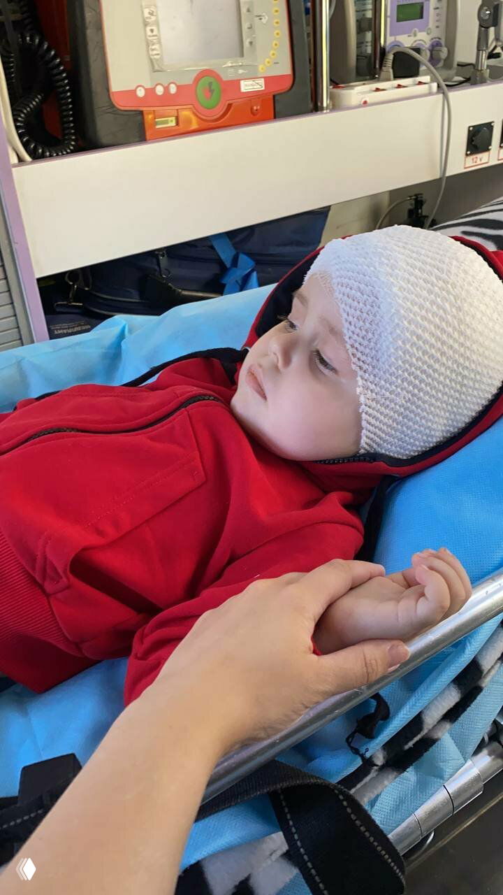 Ребёнок с перевязанной головой лежит на носилках скорой помощи, взрослый держит его руку — момент транспортировки в больнице.