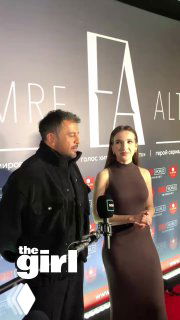 Эмре Алтуг беседует с ведущей на красной дорожке; видно пресс‑стену мероприятия, микрофон и логотип «the girl» в углу.