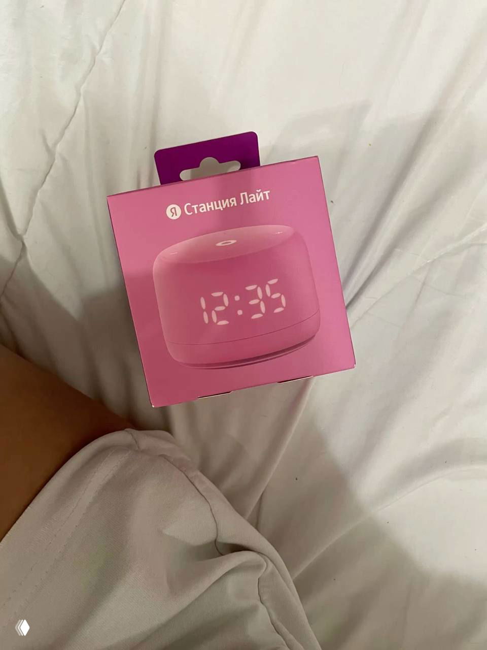 Розовая коробка с Яндекс Станция Лайт лежит на постели рядом с ногами — упаковка умной колонки как идея подарка от канала