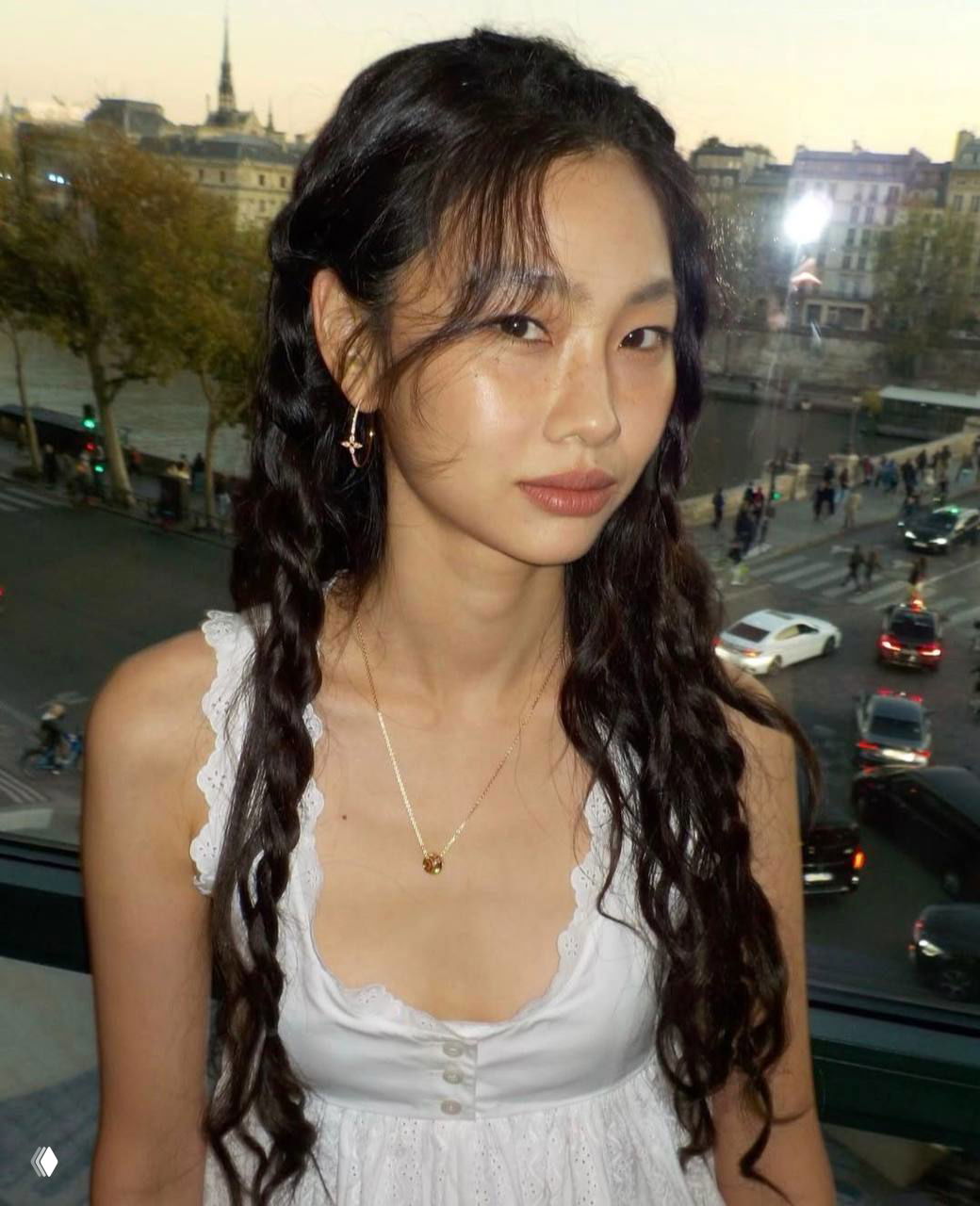 Портрет молодой азиатской женщины в белом топе на фоне городского пейзажа; естественный макияж, сияющая кожа и мягкие блики.