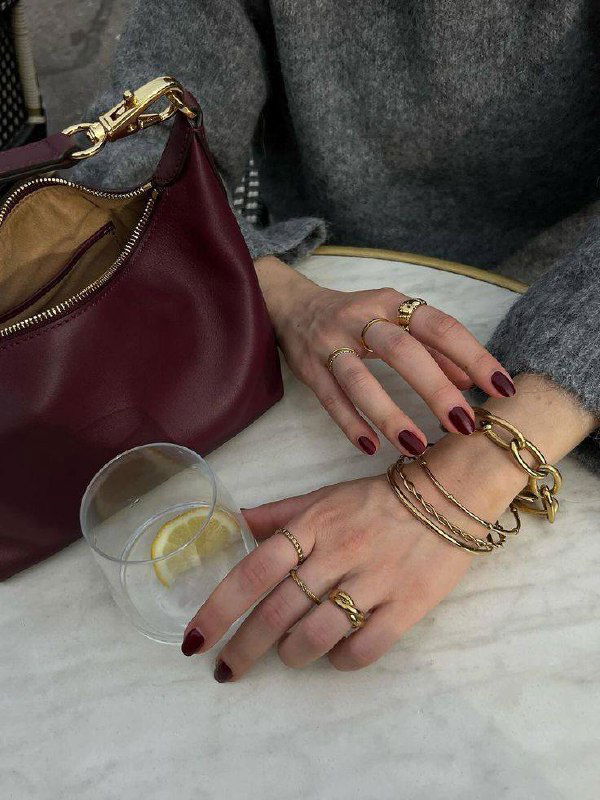 Две руки с бордовым глянцевым маникюром рядом с кожаной сумкой схожего оттенка и бокалом с долькой лимона; видны золотые кольца и браслеты, образ в кофейном кафе.