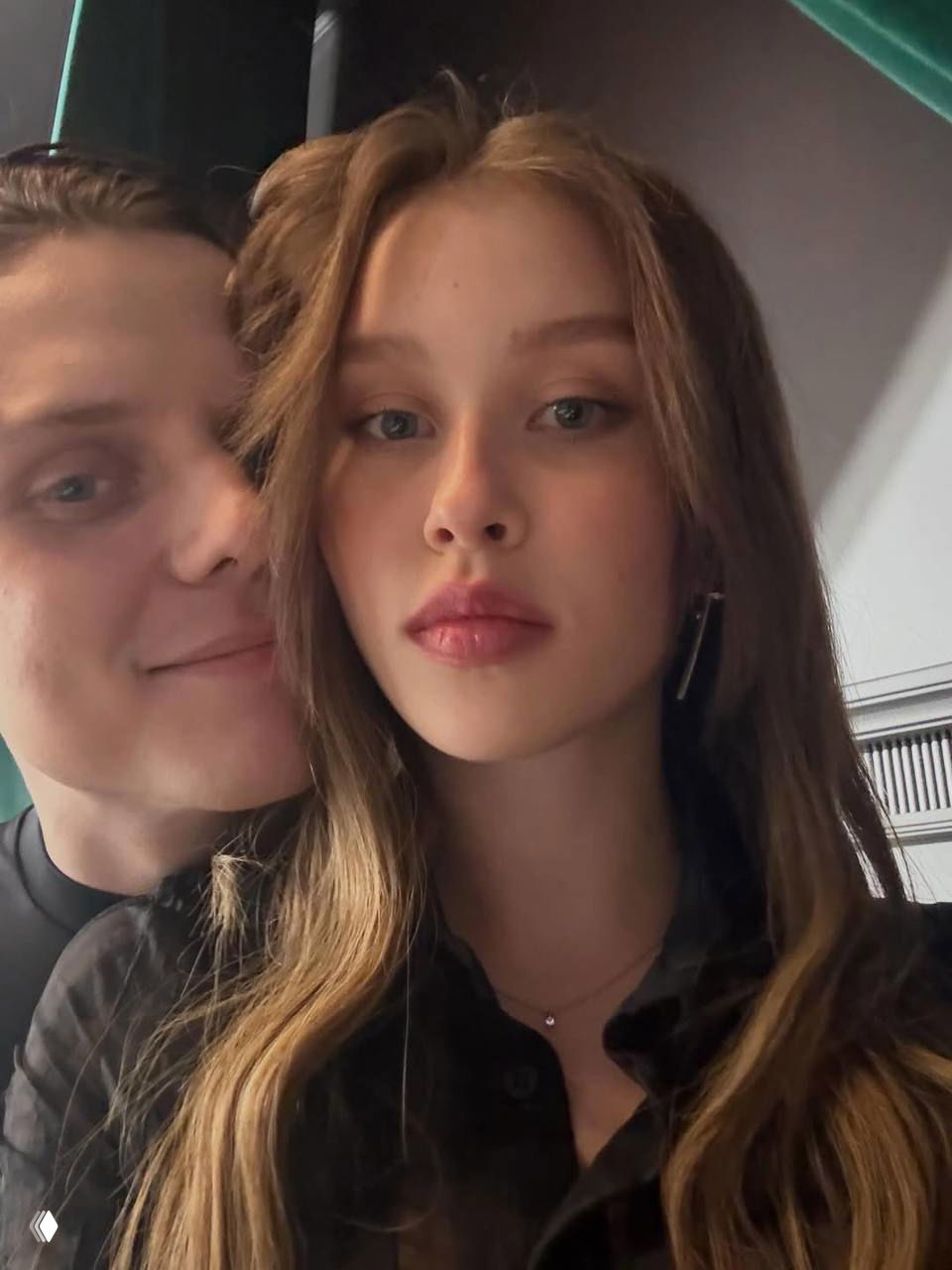 Крупный план пары на селфи: молодая женщина с распущенными светлыми волосами в центре кадра, молодой мужчина улыбается рядом на нейтральном фоне интерьера.