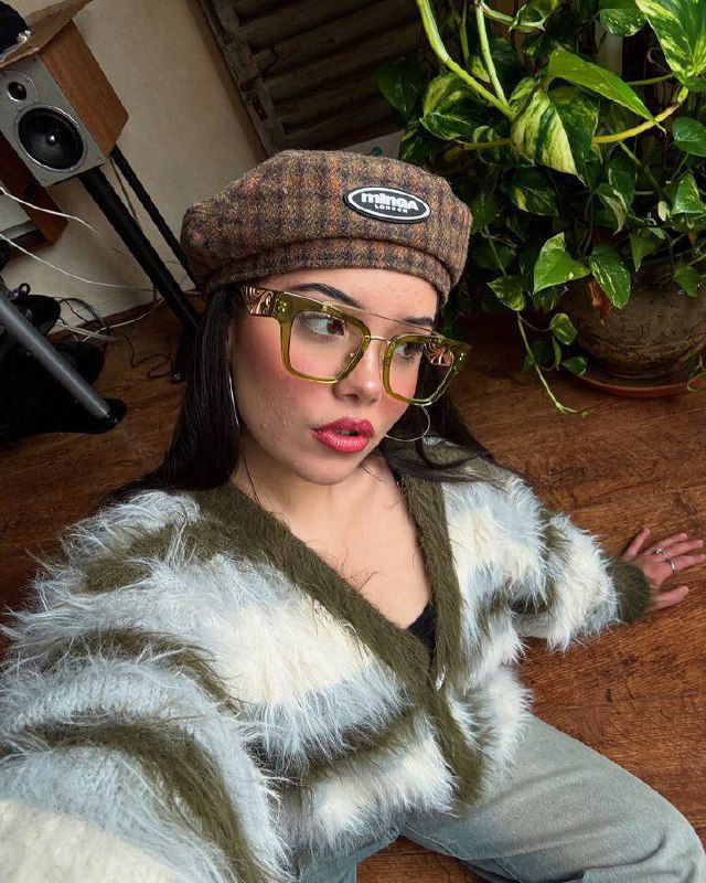 Крупный план: девушка в берете и пушистом кардигане носит крупные зелёные квадратные очки — винтажный модный образ в домашней обстановке.