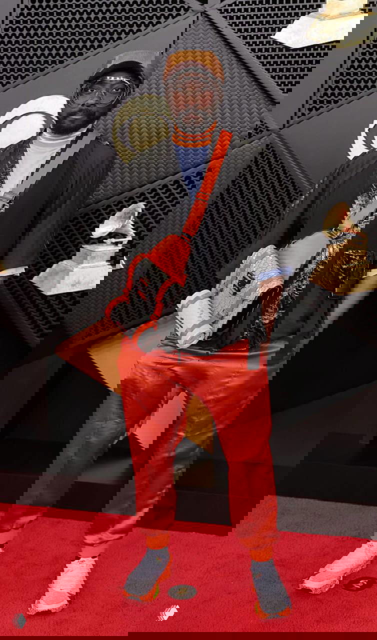 Мужчина в ярких оранжевых брюках и многослойных аксессуарах на красной дорожке «Грэмми», кроссовки, сумка через плечо и заметные очки.