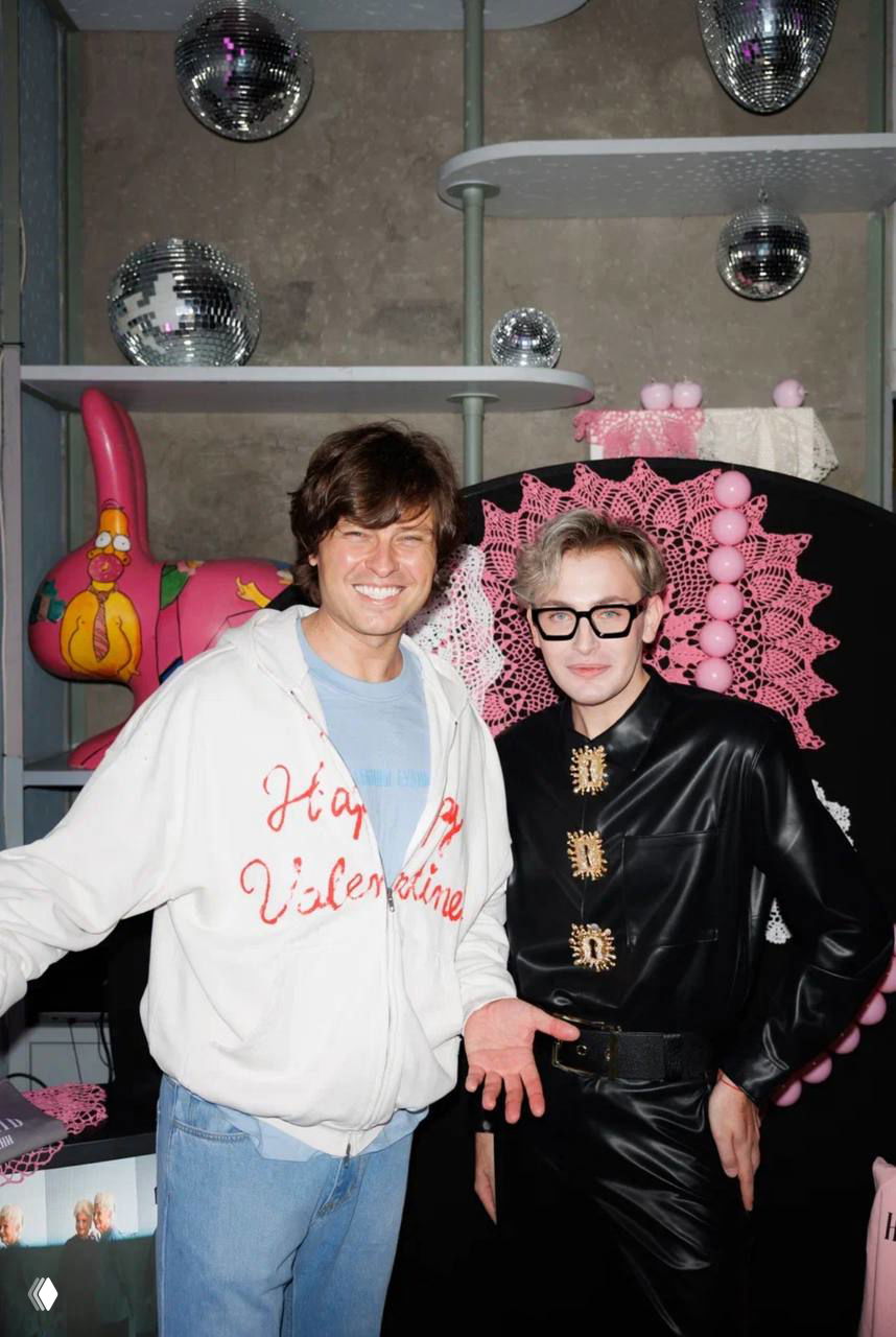 Два человека у интерьерной фотозоны с розовой вязной декорацией и диско-шарами; один в светлой толстовке, другой в чёрном кожаном комплекте на фоне сцены.