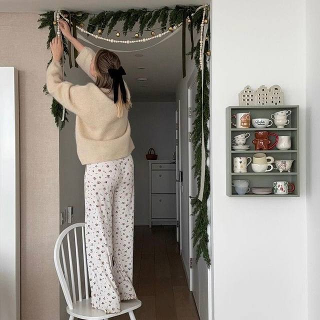 Девушка в уютном свитере и пижамных брюках вешает гирлянду на дверной проём; в кадре полка с чашками, пастельные тона и домашняя атмосфера.